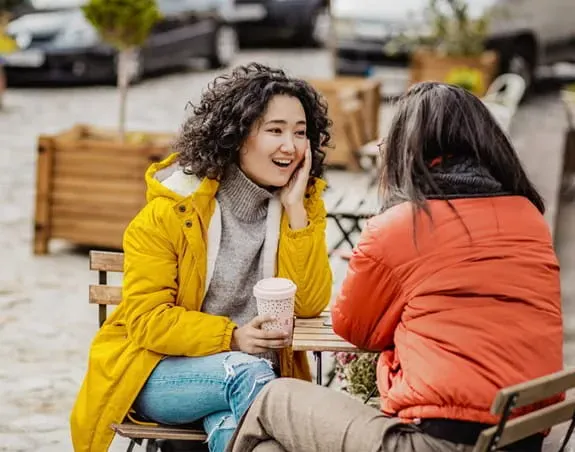 Two women in jackets sitting outside at a table with a warm beverage talking