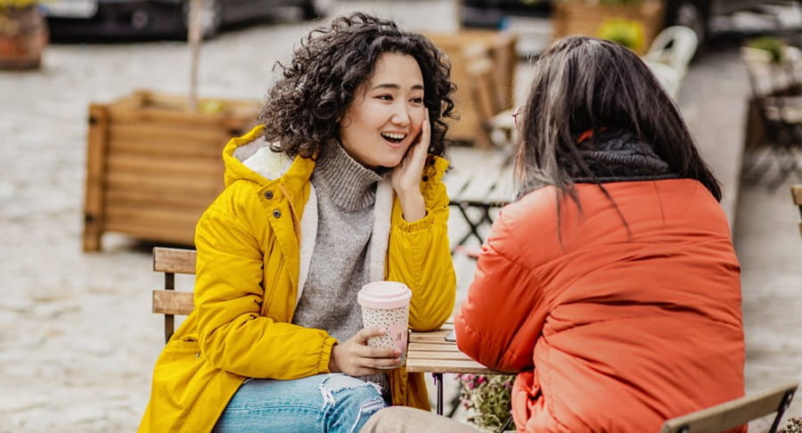 Two women in jackets sitting outside at a table with a warm beverage talking