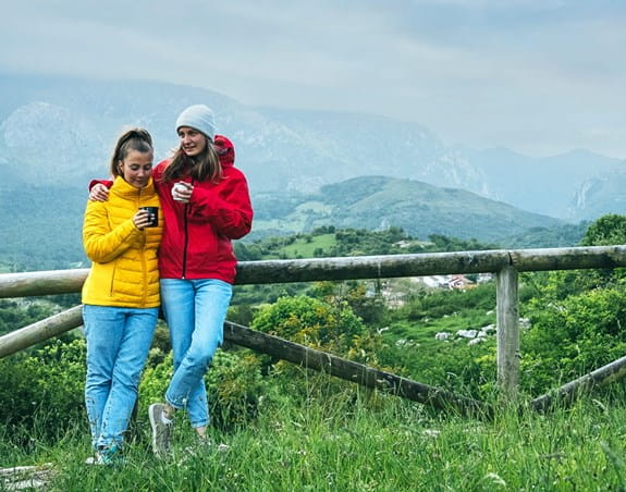 Two women in winter coats standing in a field against a wooden fence with a mountain in the distance.