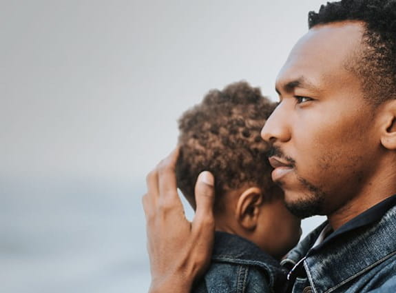 A man holds his young son’s head close to him and looks to the distance. 