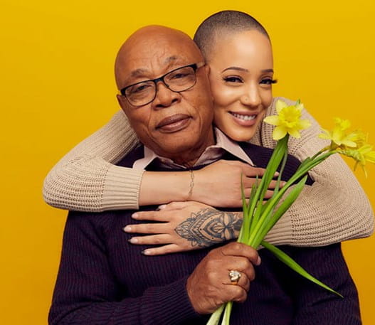 A man holds a bouquet of daffodils while a young woman embraces him over his shoulders.