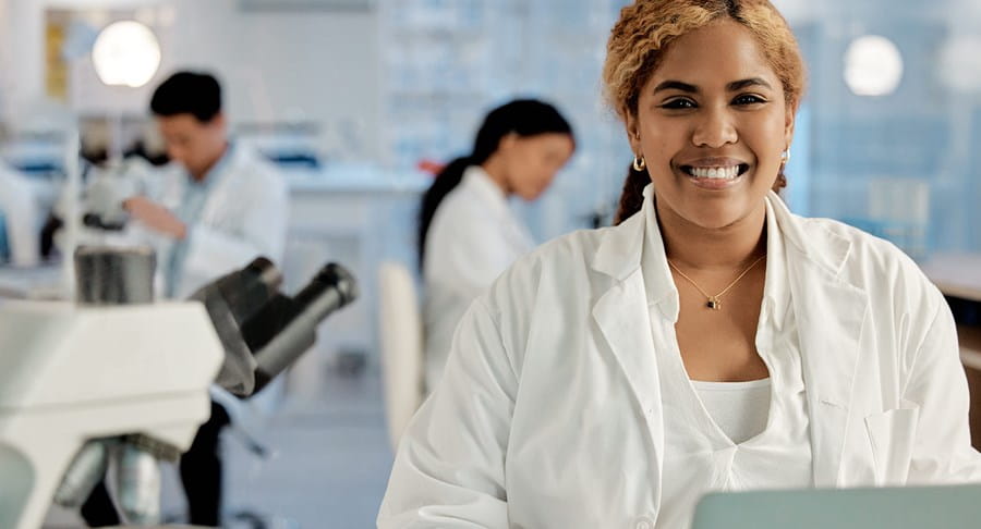 Three people in white lab coats working in a lab. One person is smiling at the camera.
