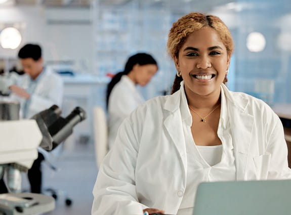 Three people in white lab coats working in a lab. One person is smiling at the camera.