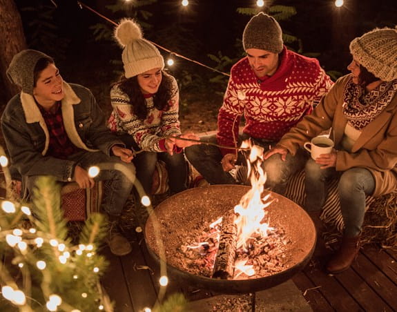 A group of people sitting around a campfire, some sipping hot chocolate and roasting marshmallows.