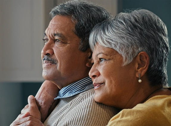 A woman embraces her husband and rests her chin on his shoulder while they look into the distance.