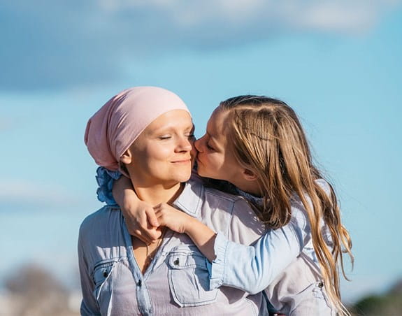 A child wraps her arms on her mother’s shoulders and kisses her on the cheek.