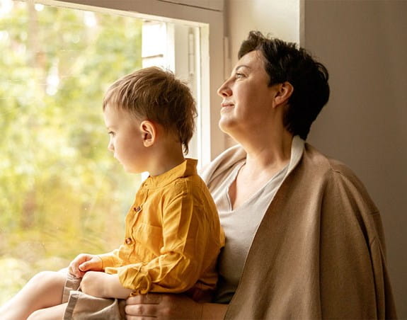 A mother holds her child while they both look out a window.