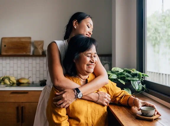 A young woman hugs an older woman from behind in a kitchen as the older woman sits by a window holding a cup and smiling.