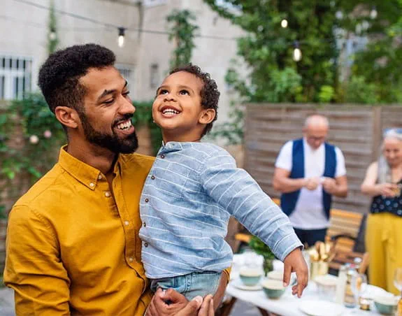 Un homme portant une chemise jaune tient un enfant souriant lors d’un rassemblement en plein air. Deux adultes sont en arrière-plan et préparent de la nourriture et des boissons.