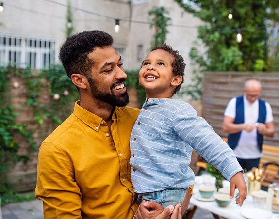 A man in a yellow shirt holds a smiling child at an outdoor gathering, with two adults in the background preparing food and drinks.