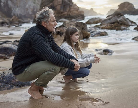 Grandfather and granddaughter crouching at the beach looking at view