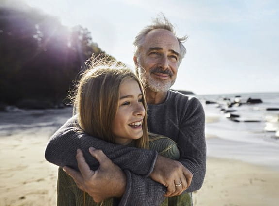 An older man stands on the beach with his arms wrapped around his daughter. They both stare out into the distance.