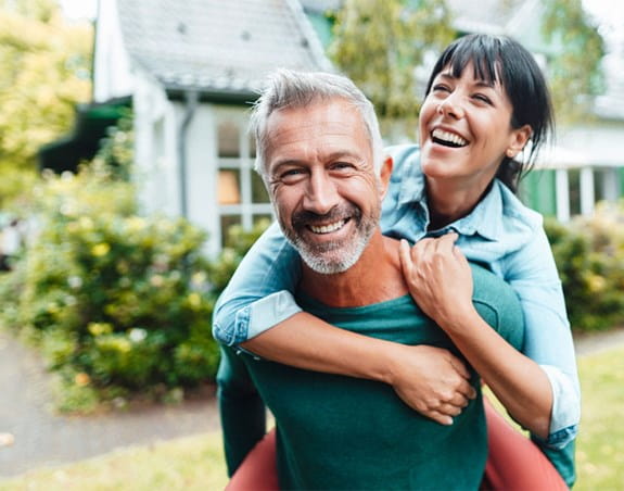 A woman is hanging onto the back of a man playfully. The man stares into the camera while the woman stares off into the distance. They are both smiling.
