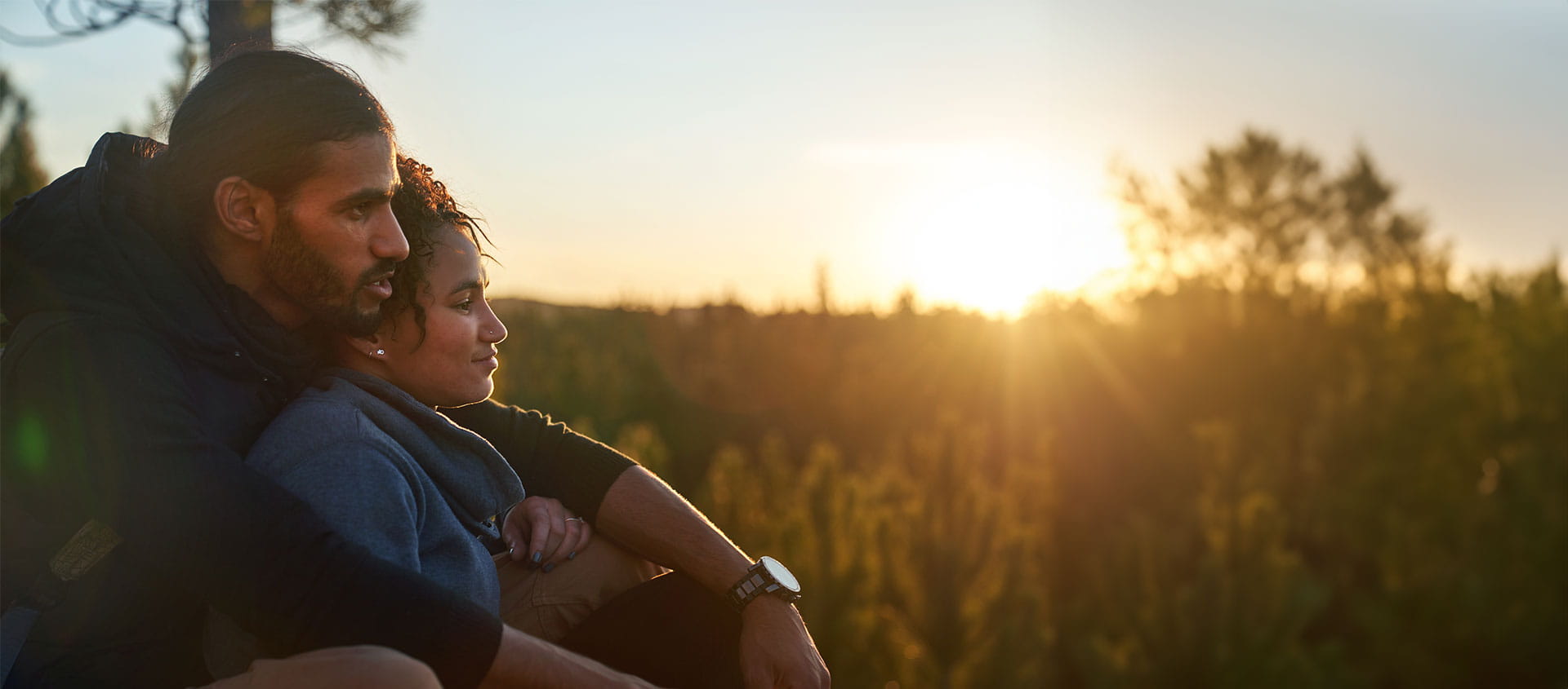 A young couple in close proximity watching the sun setting on a hill top.  