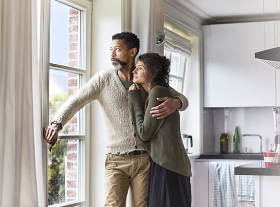 A man stands looking out a window with his arm wrapped around a woman. She also stares out the window.