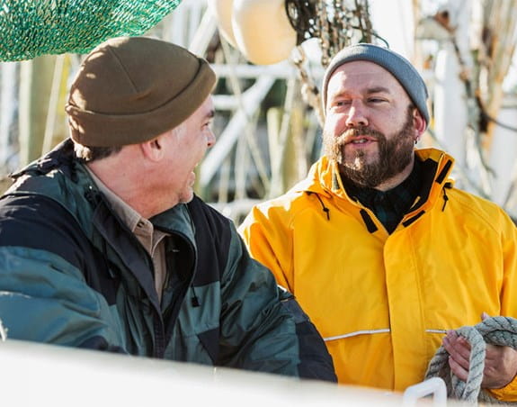 Deux hommes travaillent sur un bateau, habillés pour le temps pluvieux avec des tuques et des vestes imperméables. L’un d’eux rit.
