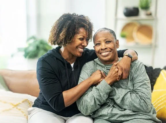 A woman sits on a couch with her arms around an older woman. They are both smiling.