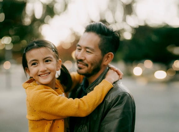 A man stands smiling looking at his daughter. She has her arms wrapped around his shoulders as he holds her. She smiles off into the distance.