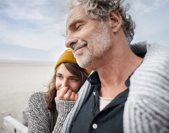 A man stands with his eyes closed on the beach. A woman stands beside him leaning on his shoulder with her hand over her mouth.