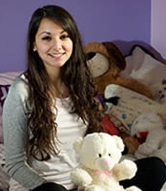Girl with long brown hair sitting on her bed smiling
