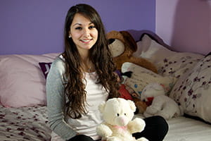 Girl with long brown hair sitting on her bed smiling