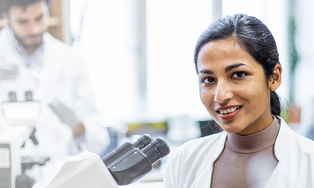Une chercheuse assise à côté d’un microscope souriant à l’objectif.