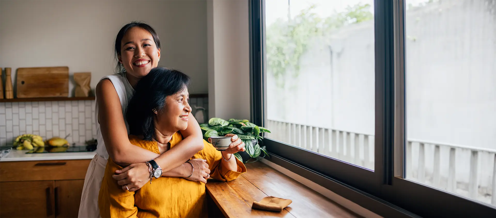 Une jeune femme enlace une femme plus âgée par derrière dans une cuisine alors que la femme plus âgée est assise près d’une fenêtre, tenant une tasse et souriant.