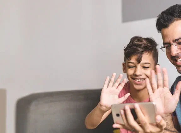 A father wearing glasses, smiling with a child sitting on a couch waving to a cell phone.