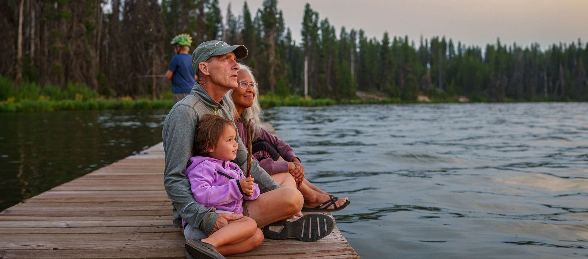 Des grands-parents et leur petit-enfant assis sur un quai au bord d'un lac, en pleine nature.