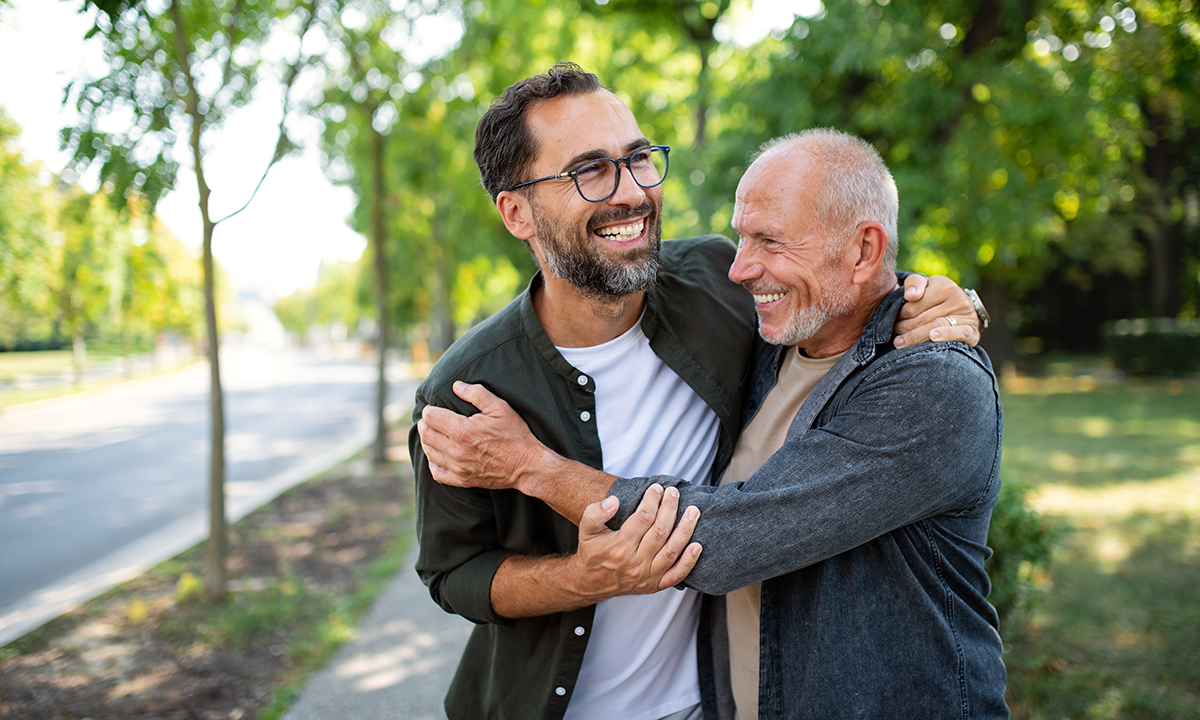 An older and a younger person walk together outdoors with their arms around each other on a tree-lined path.