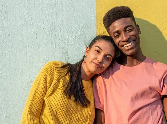 A young man and woman stand shoulder to shoulder in front of a colourful wall.