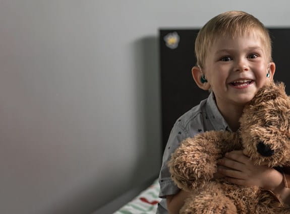 Young boy holding a brown teddy bear, sitting on his bed, smiling
