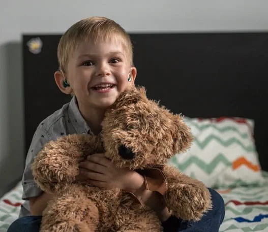 Young boy holding a brown teddy bear, sitting on his bed, smiling
