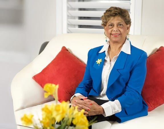 Woman with short, brown hair, sitting on a couch, wearing a blue blazer, yellow daffodil pin, white collared shirt, smiling