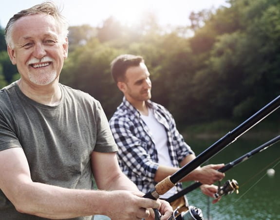 Older man, facing forward, smiling with younger man, fishing.