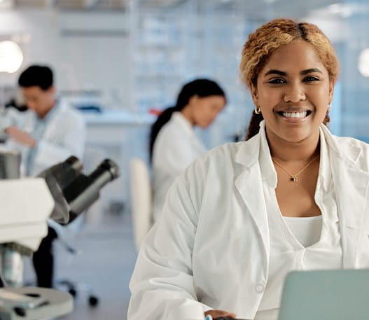 Three people in white lab coats working in a lab. One person is smiling at the camera.